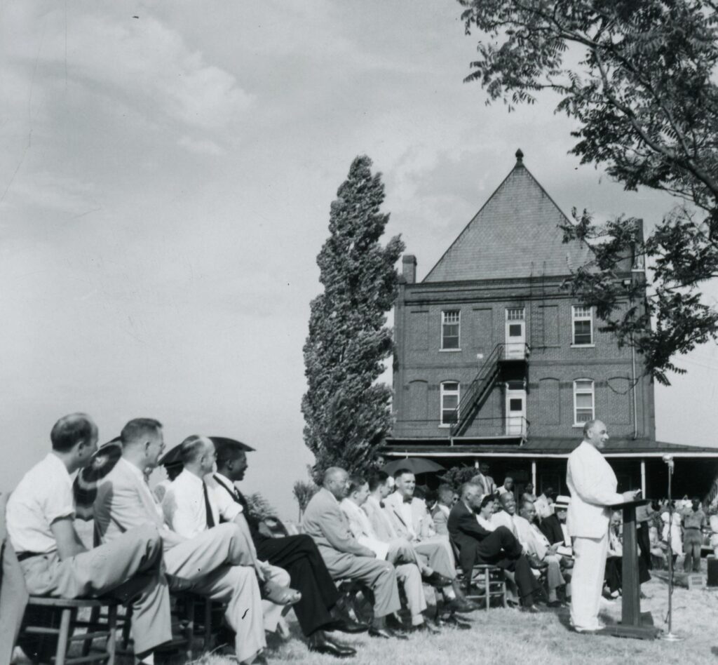 A large crowd is pictured behind a man speaking at a podium with the side of Burrell Hospital in the background.