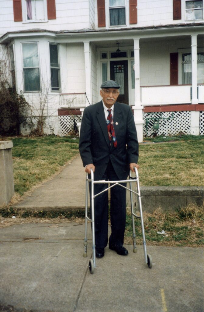 Oliver Hill standing in a dapper suit and hat in front of his childhood home, with white paint and red shutters, in Gainsboro