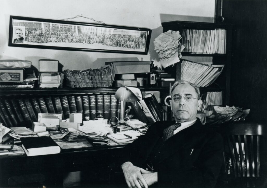 The Doctor sitting in his office filled with books and a long photo in the background